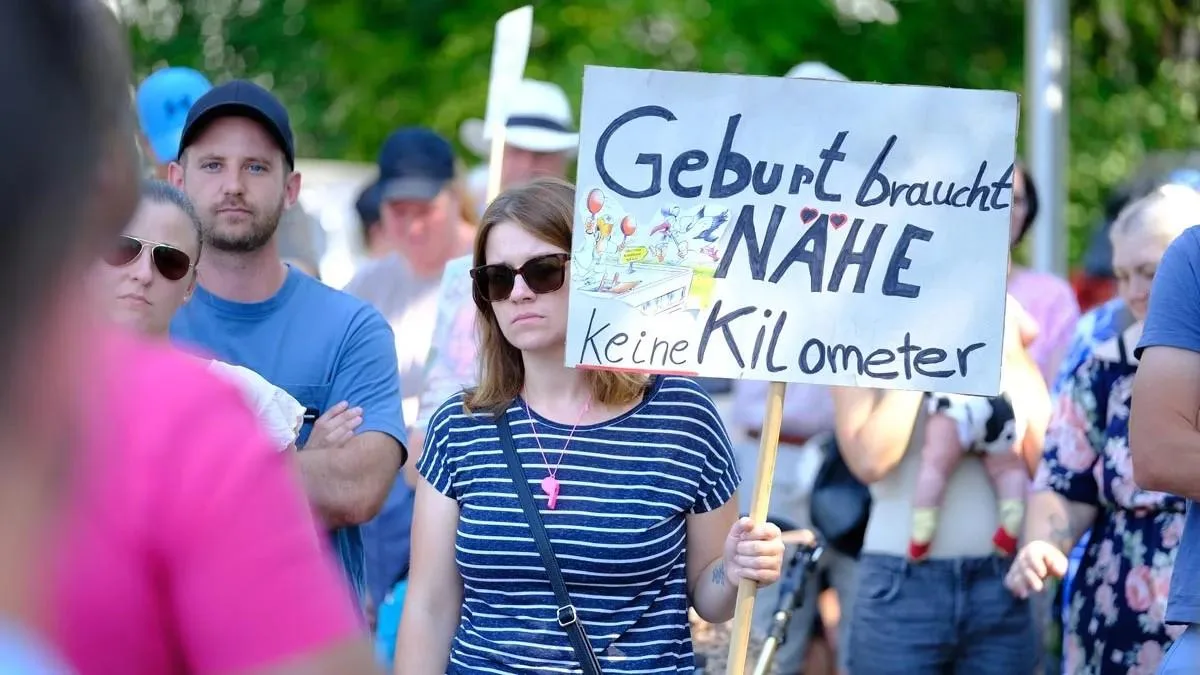 Ein Schild mit der handgeschriebenen Aufschrift „Geburt braucht Nähe. Keine Kilometer.“ wird in die Höhe gehalten.