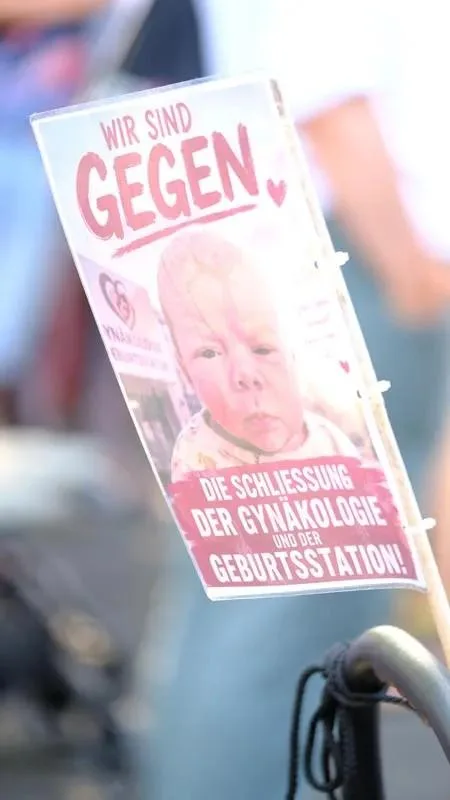 Gedrucktes rosafarbenes Plakat mit einem Babyfoto und der Aufschrift „Wir sind gegen die Schließung der Gynäkologie und Geburtsstation“.